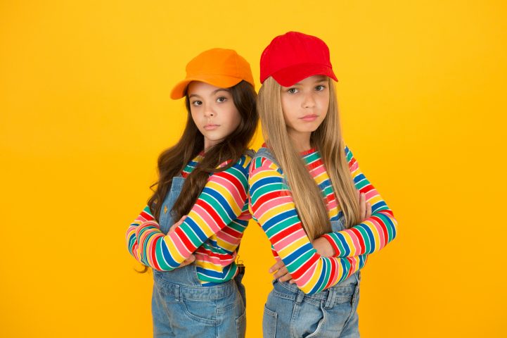 Fashion that matches their conscious style. Small girls in style keeping arms crossed on yellow background. Little children wearing baseball caps in casual streetwear style. Hip hop or hipster style.