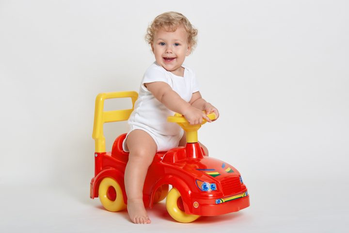 Charming baby boy sitting on red and yellow tolocar, wearing white body suit, has blond curly hair, cute infant looking directly at camera with excited facial expression.